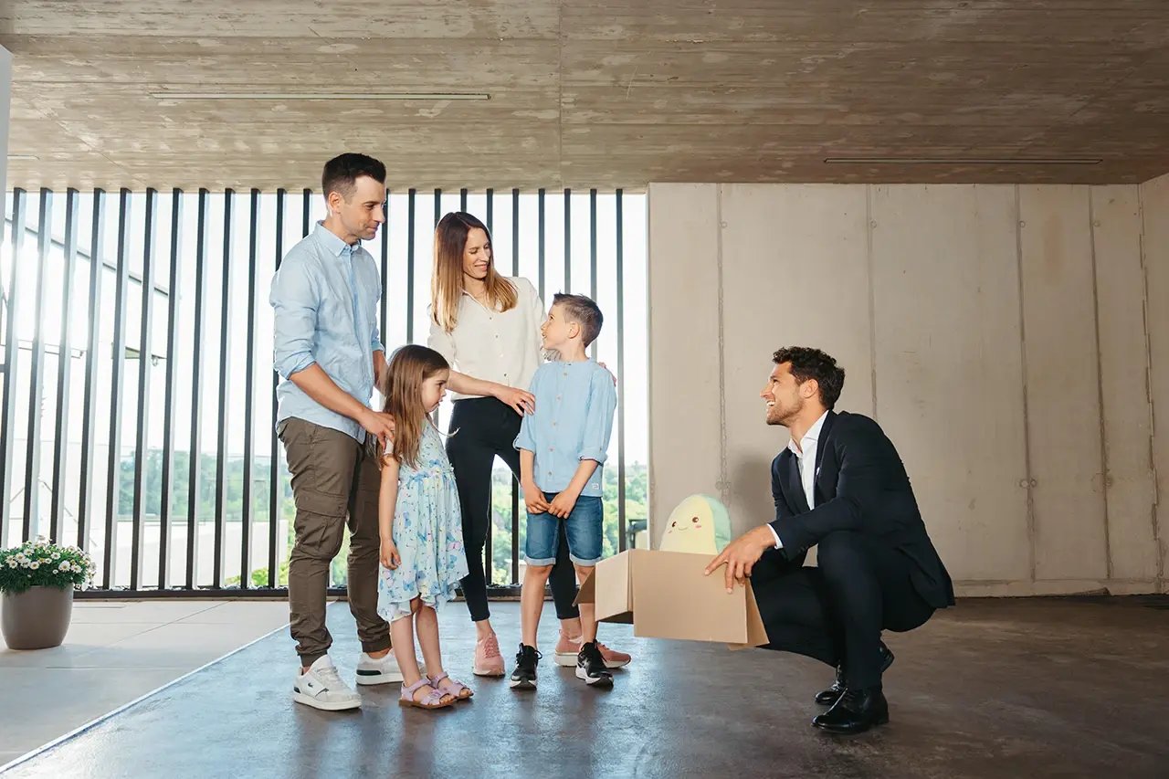 Agente immobiliare accompagna una famiglia con due bambini nella visita di una nuova casa, con uno scatolone e un peluche simbolo del trasloco.